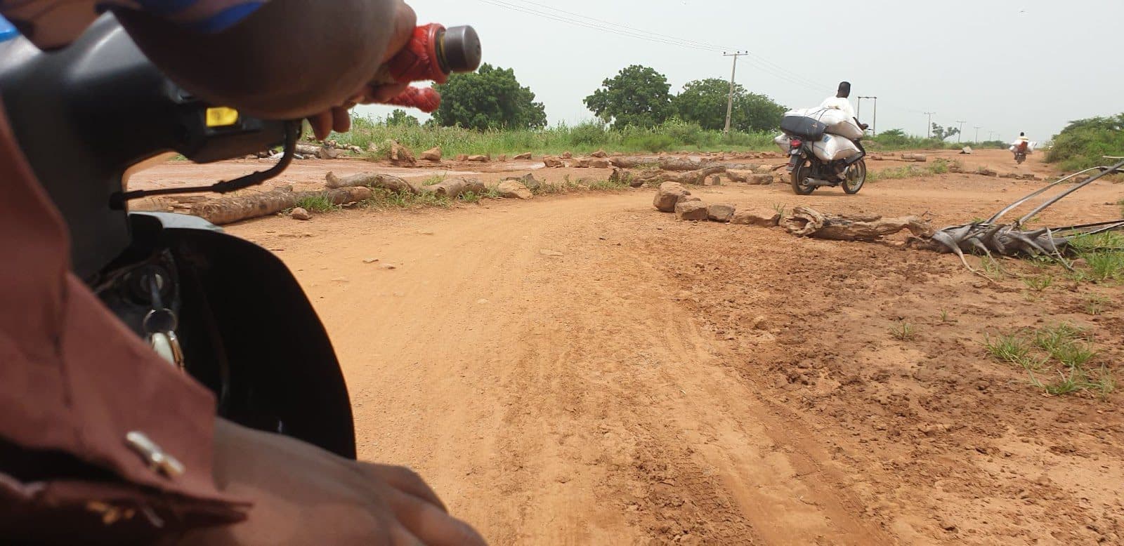 First-person view from a motorcycle approaching a dirt road intersection with a passing rider and scattered rocks.