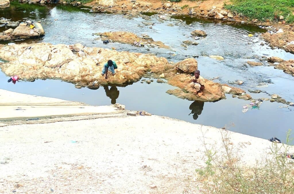 Two people sitting on rocks by a stream with litter visible in the water and on the banks.