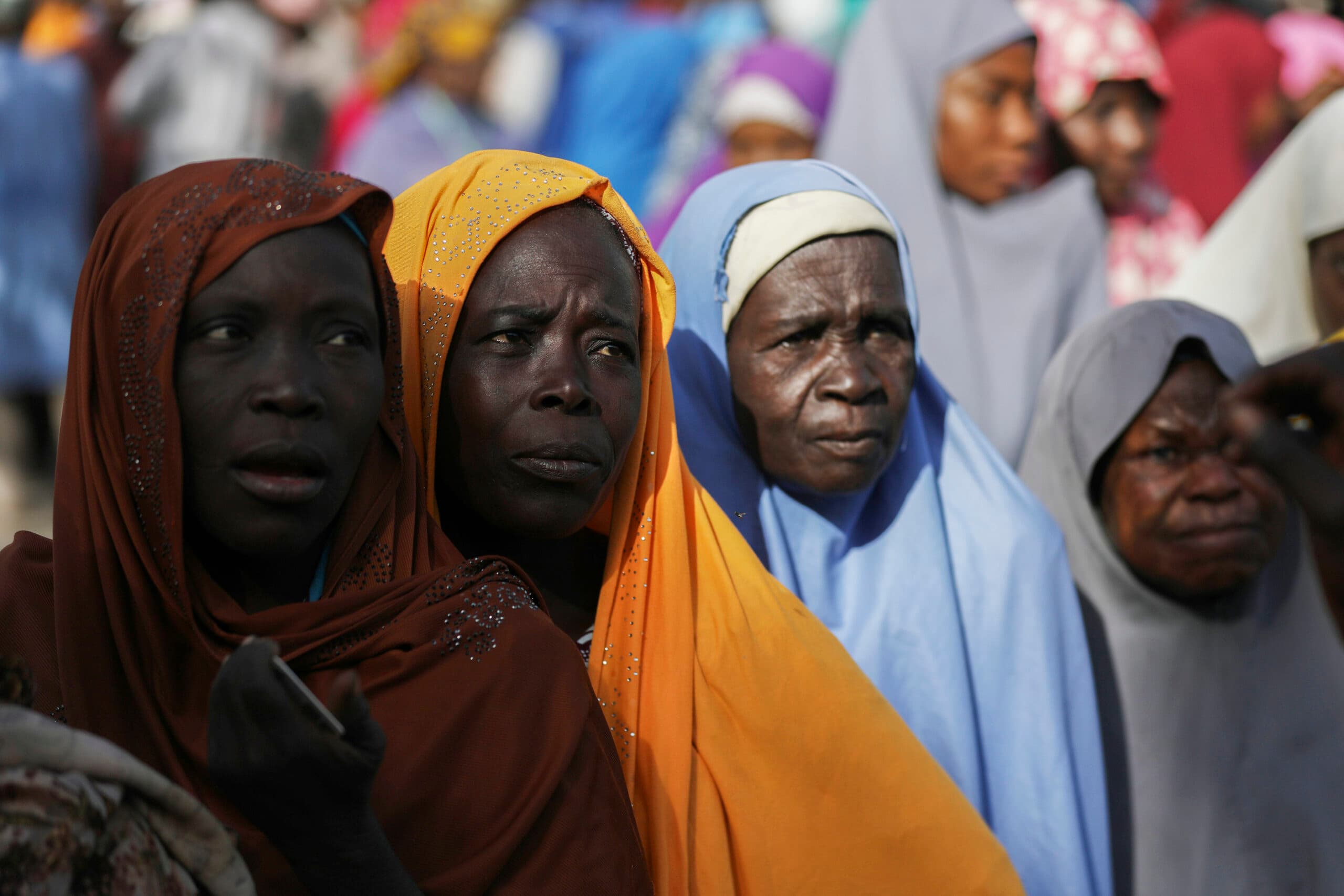 Group of women with contemplative expressions wearing colorful headscarves in a crowd.