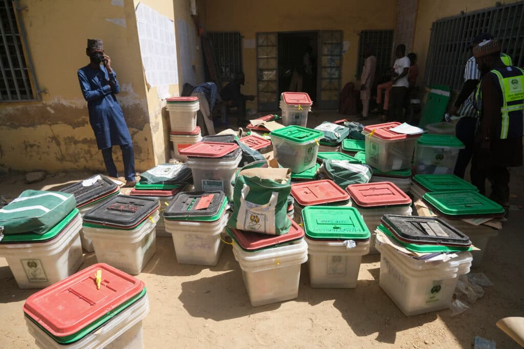 A collection of stacked ballot boxes outside a building with observers and officials present during an election.