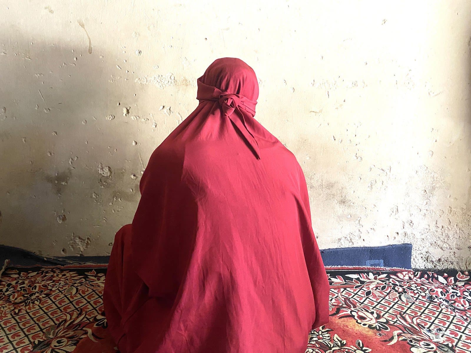 Person in red cloak sitting against a weathered wall on a patterned carpet.