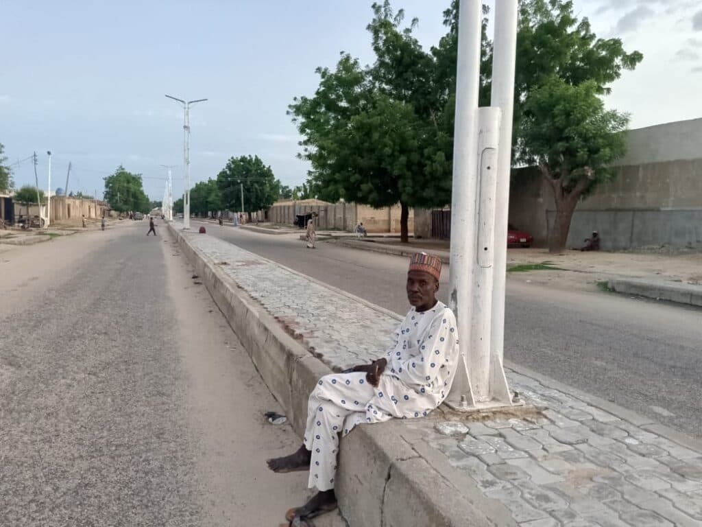 Man in traditional attire sitting on a curb by a street with trees and light poles.