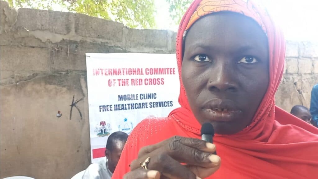 A woman in a red headscarf holding a microphone, with a Red Cross mobile clinic banner in the background.