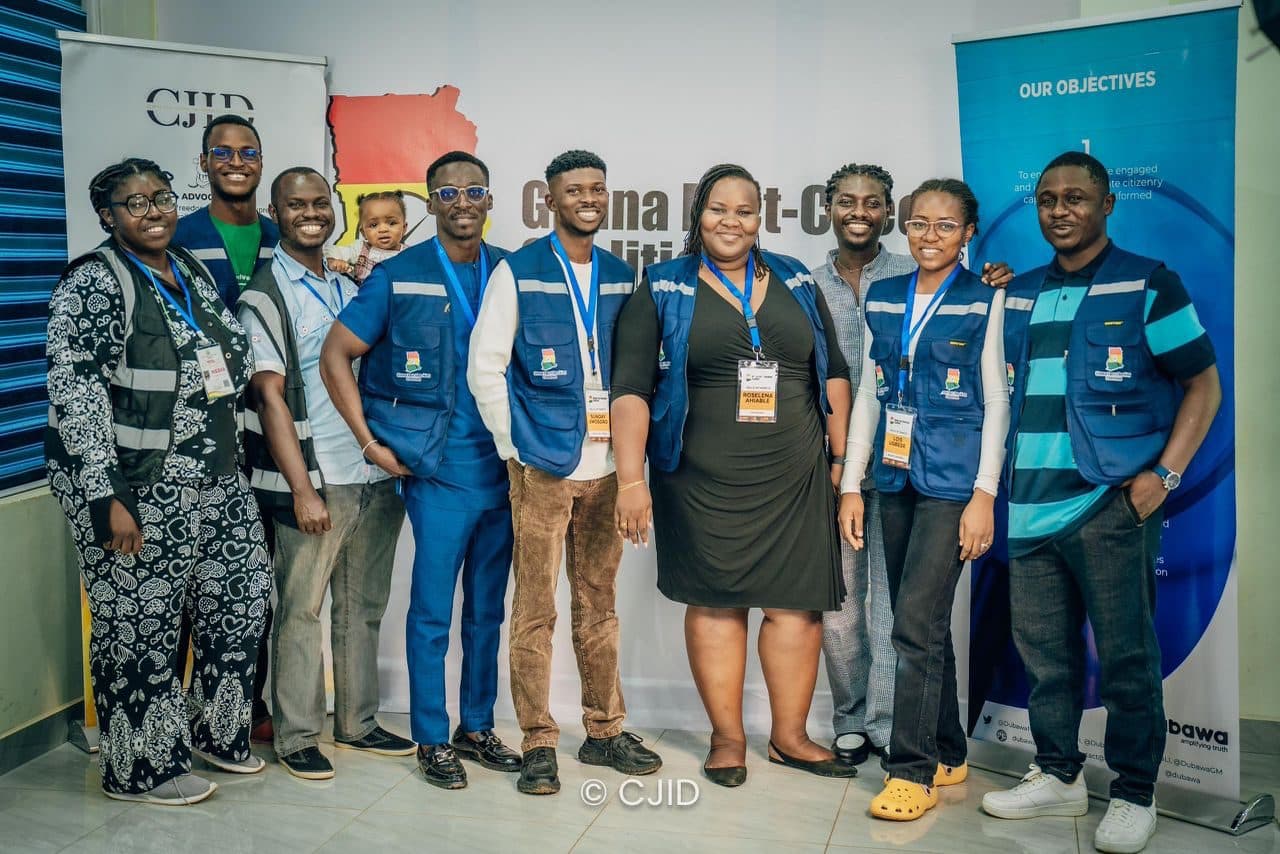 Group of smiling people wearing matching vests and ID badges in front of a "Ghana Fact-Check" banner.
