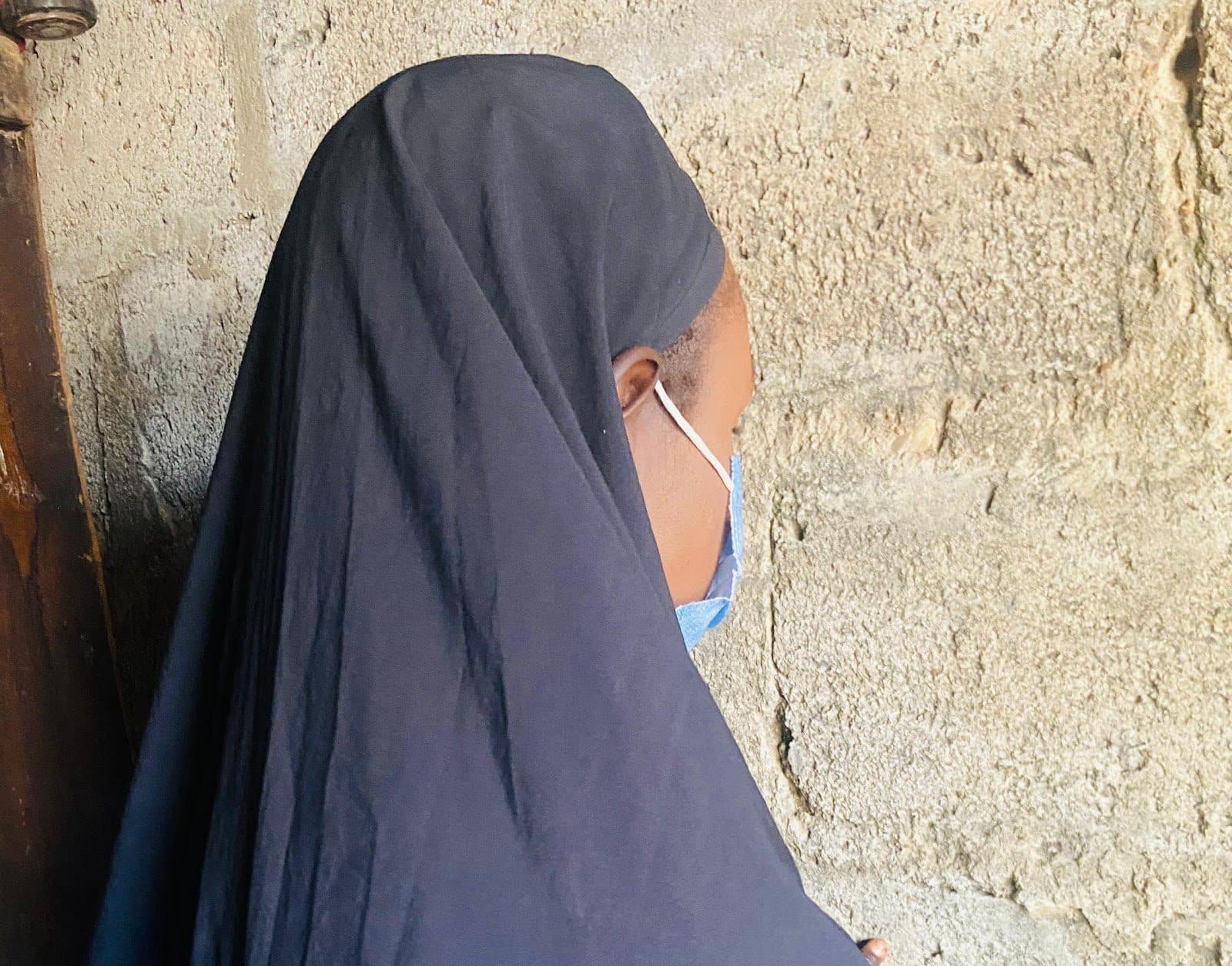 Person in a dark veil leaning against a textured wall, partially facing away.