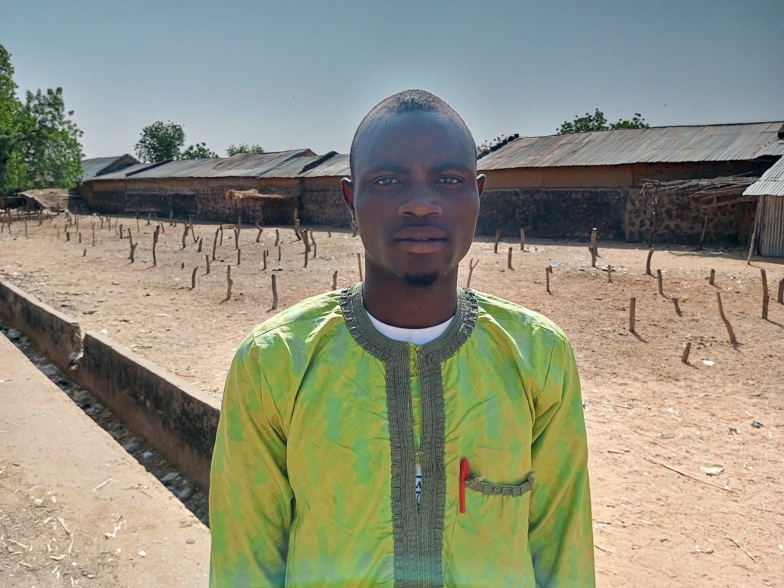 Man in a green patterned shirt standing outdoors on a sunny day, with rustic buildings and trees in the background.