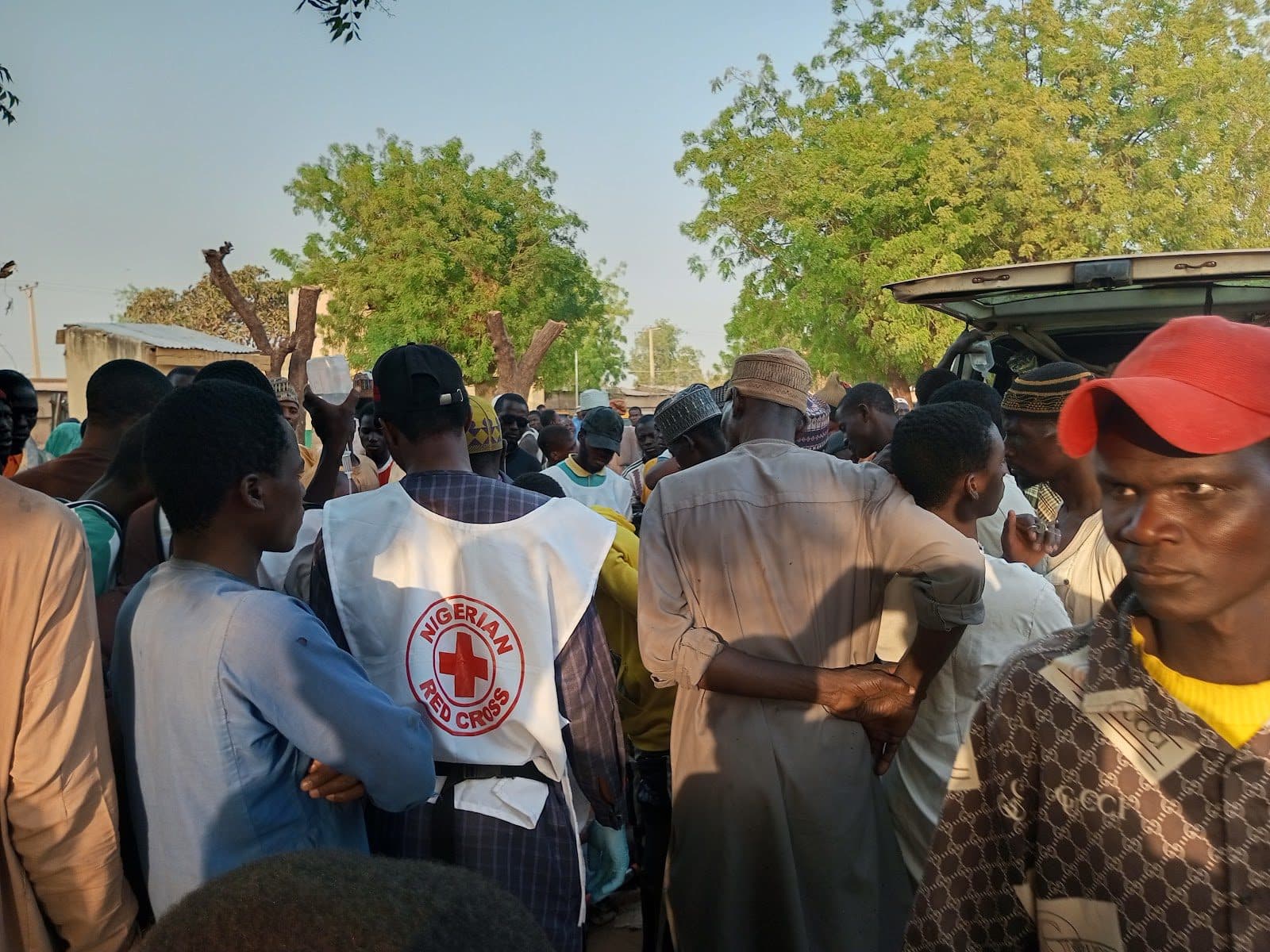 A group of people gathered around a Nigerian Red Cross worker outdoors, with trees and a vehicle in the background.