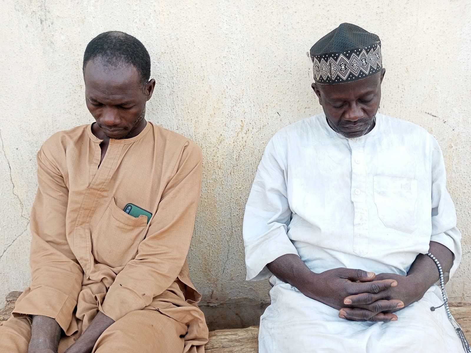 Two men, one in beige and the other in white with a cap, sit side by side against a textured wall, heads bowed, appearing contemplative.