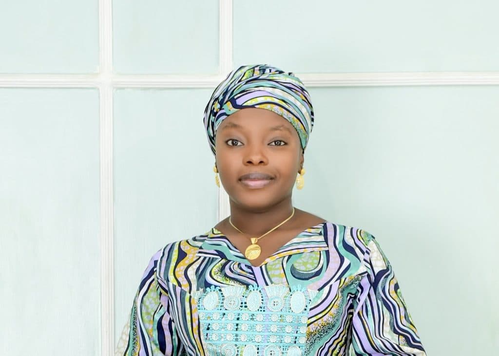 Woman in colorful patterned attire and headscarf, with a calm expression, standing in front of a light-colored wall.