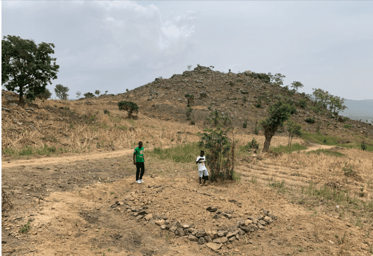Two people stand on a rocky path in a dry, hilly landscape with scattered trees under a cloudy sky.