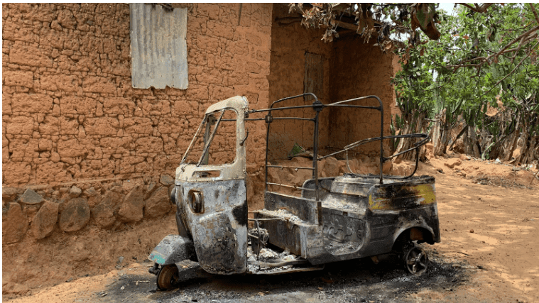 Burnt-out tuk-tuk with missing parts, parked beside a mud-brick building, surrounded by trees.