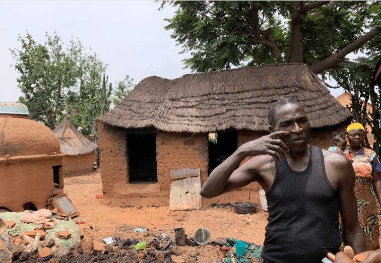 Man in front of a thatched-roof hut, gesturing with his hand. Others stand nearby, with trees and rural setting in the background.