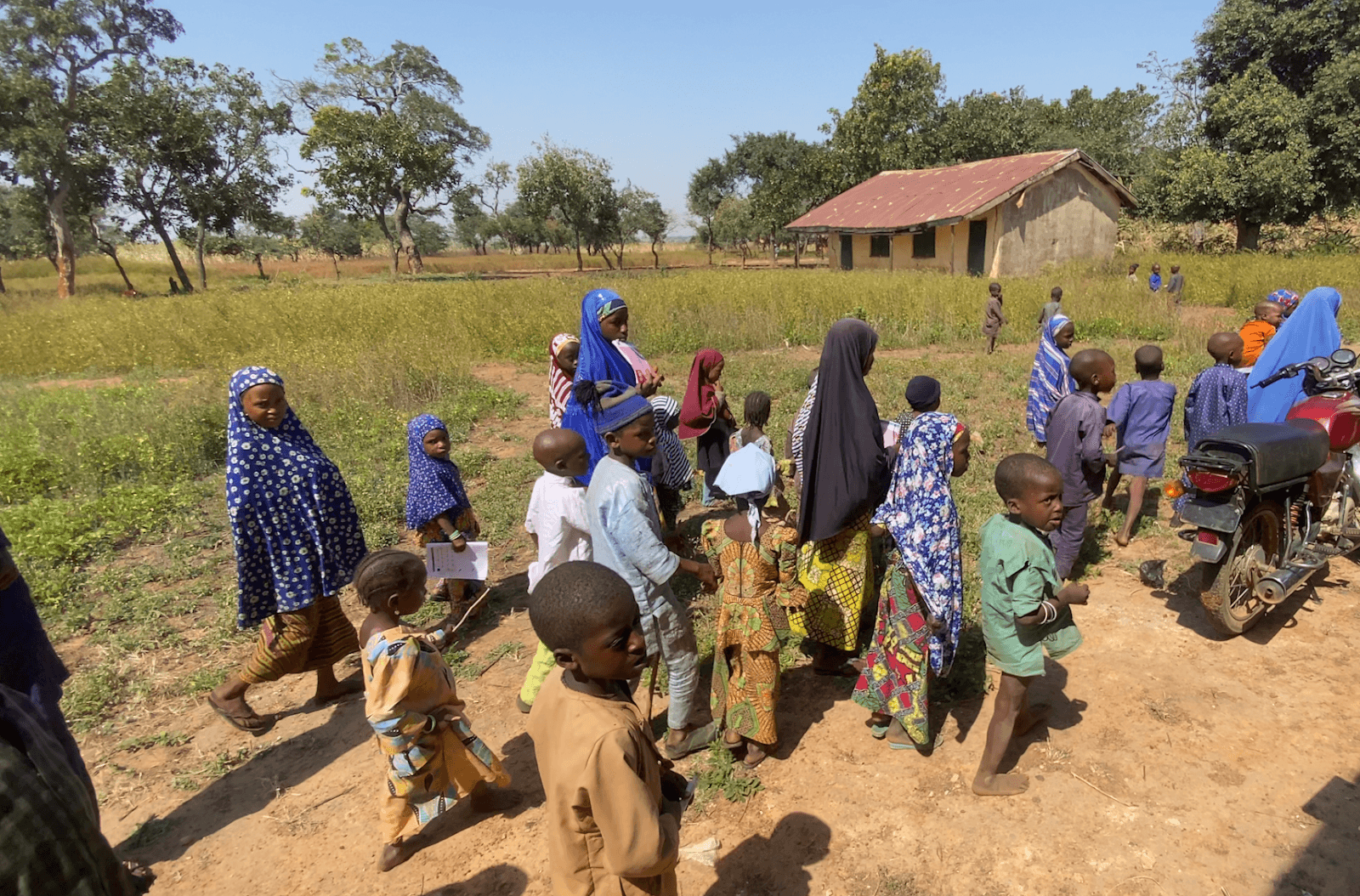 A group of children walks outdoors near a small building surrounded by grass and trees, under a clear blue sky.