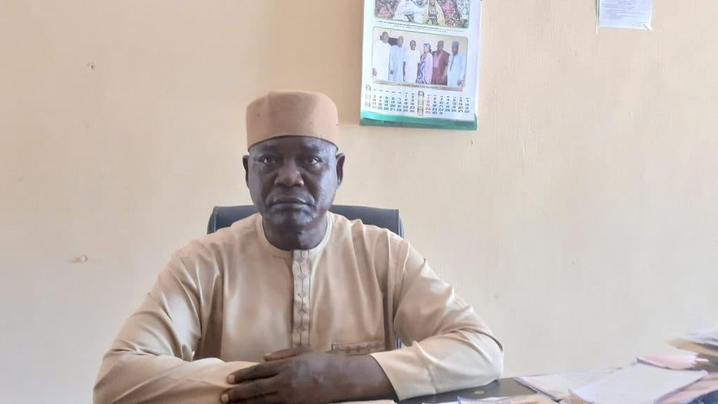 Man in traditional attire sitting at a desk with hands folded, a calendar on the wall behind him.