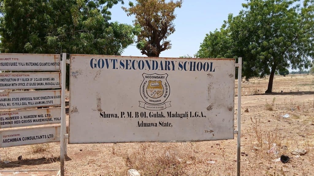 Sign for Govt Secondary School, Shuwa, Adamawa State, Nigeria, with project details on adjacent board, surrounded by trees.