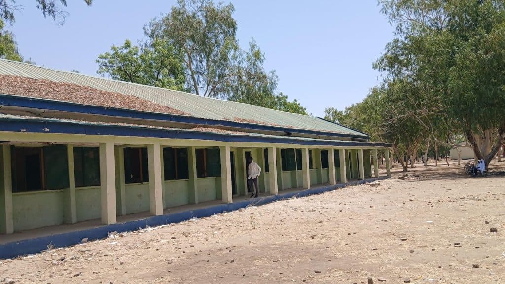 A person stands near a long, single-story building with green walls and a tin roof, surrounded by trees and dry ground.