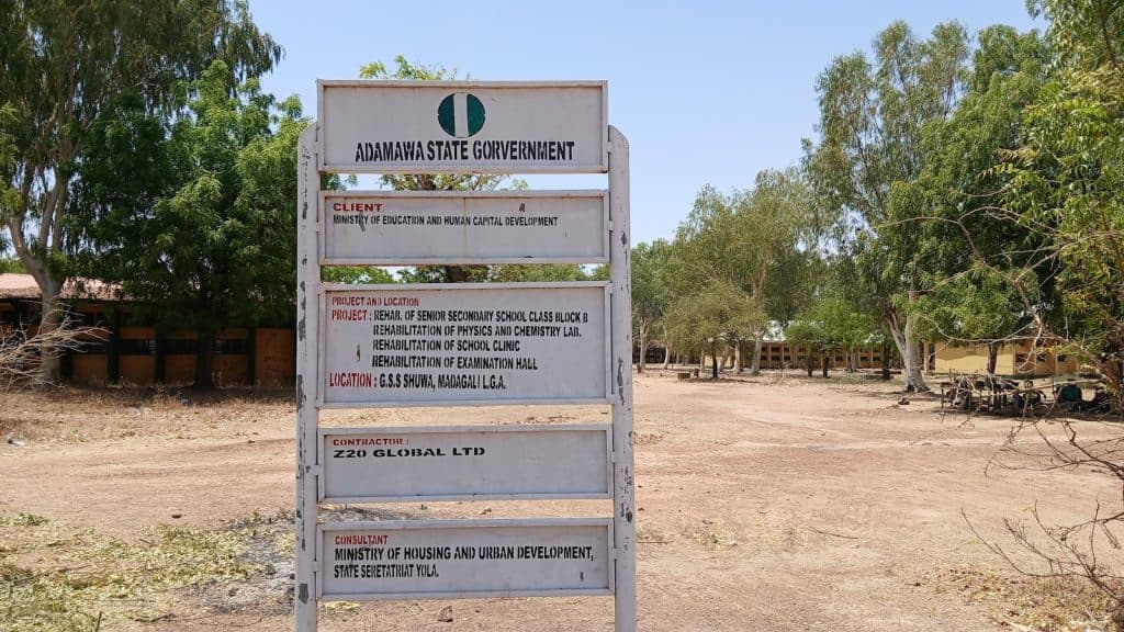 A sign in a dry, tree-lined area details a school rehabilitation project by Adamawa State Government, including labs and a clinic.