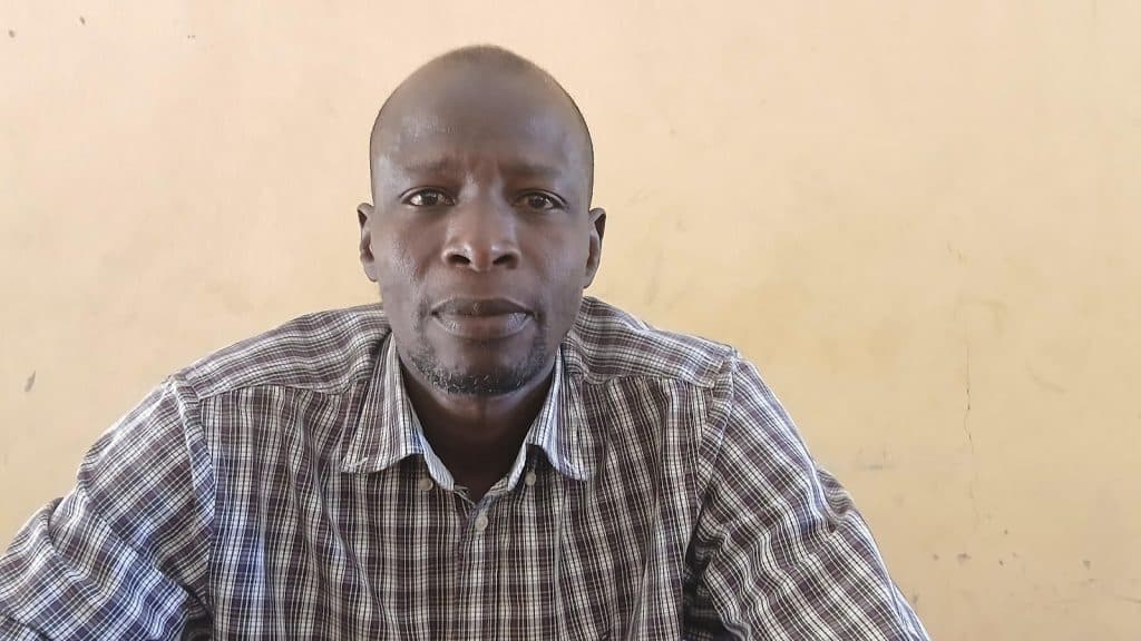 A person in a plaid shirt sits against a plain light-colored wall, looking directly at the camera.