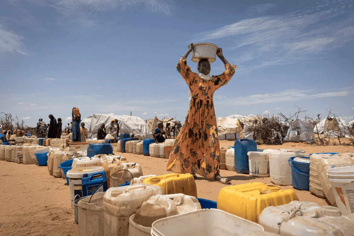A woman carries a bucket on her head near rows of water containers in a dry landscape with tents and people in the background.