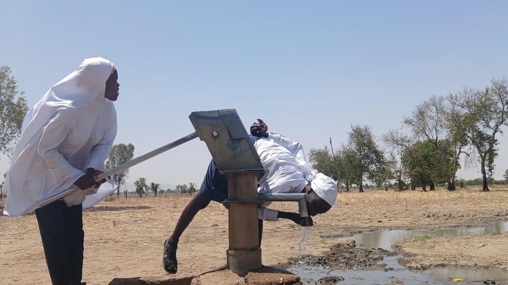 Two individuals in white clothing use a hand pump to collect water in a rural setting with trees and open sky.