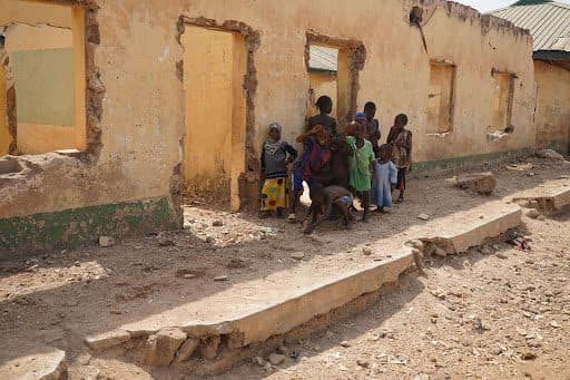A group of people stand outside a worn, roofed building with missing doors and windows in a dry, dusty environment.