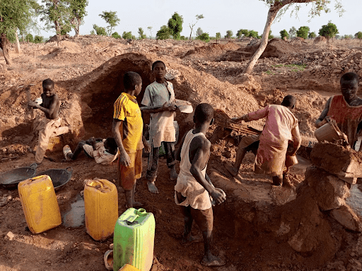 Children working in a rural area with tools and water containers, surrounded by dirt and trees.