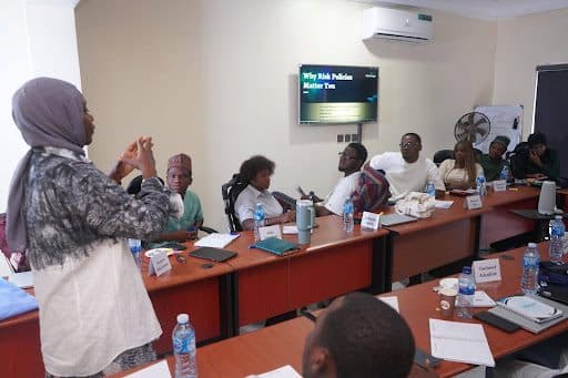 A woman presents to a seated group in a classroom with a screen displaying "Why Risk Policies Matter to...", laptops and notes visible.