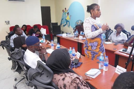 A woman is speaking in a meeting room with attendees seated at tables, listening and taking notes.