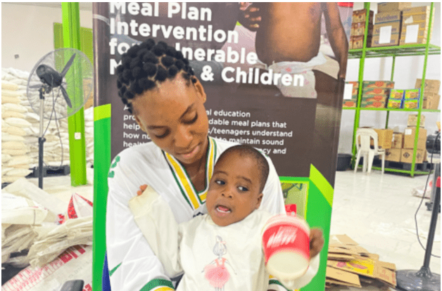 Woman holding a child in front of a "Meal Plan Intervention" poster inside a facility with shelves and bags.