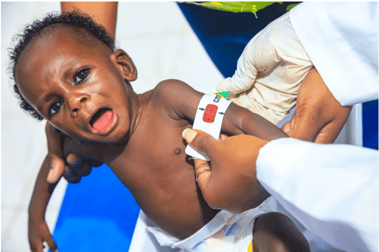A baby is being measured with a Mid-Upper Arm Circumference (MUAC) tape by a gloved healthcare worker.