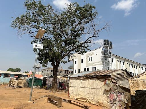 A large tree beside a solar streetlight, near worn structures, with a modern white building in the background under a clear sky.
