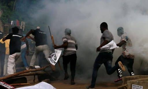 People running through smoke, carrying banners and sticks, in a chaotic scene.