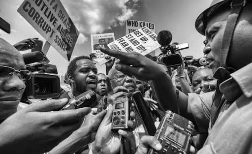 Crowd of reporters interviewing a man, surrounded by protest signs. People hold microphones and cameras in a tense atmosphere. Black and white photo.