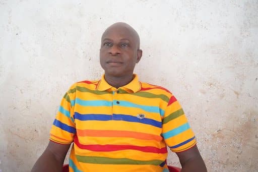 Person wearing a colorful striped shirt sits against a textured white wall, looking relaxed.