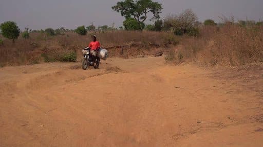 Person riding a motorcycle on a dusty rural path, surrounded by dry grass and sparse trees.