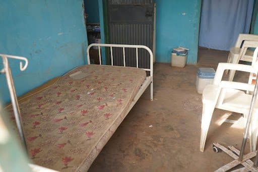 A room with a worn-out bed, plastic chairs, and peeling walls, suggesting a simple, unadorned setting.