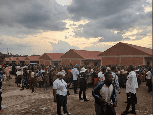 Crowd gathered outdoors near red-roofed buildings at sunset, under a cloudy sky.