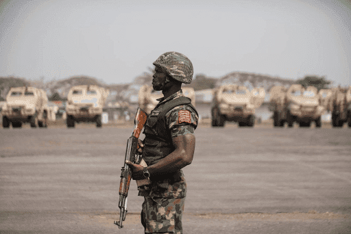 A soldier in camouflage uniform stands guard with a rifle. Armored vehicles are lined up in the background.