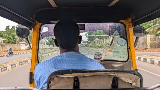 View from the back of a tuk-tuk, focusing on the driver, with a street scene visible through a cracked windshield.