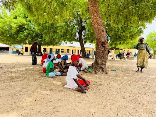 Children sitting under trees for shade on a sunny day, with a woman walking nearby and a building in the background.