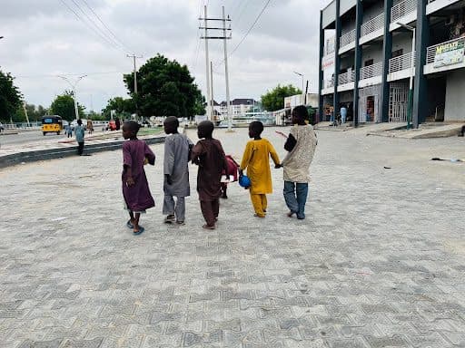 Five children walk hand in hand on a paved street near buildings, holding a blue balloon, cloudy sky above.