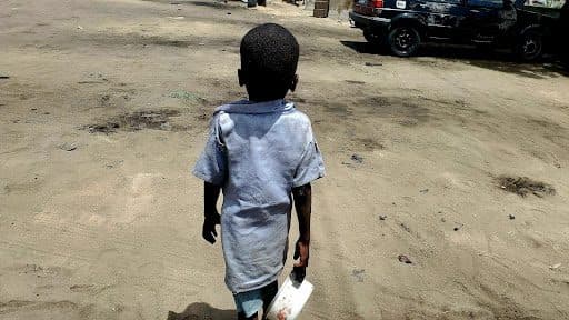 A child in a blue shirt walks on a sandy street, holding a white container. A car is parked in the background.