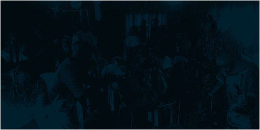 A group of people sitting and standing in a dimly lit room, some appear to be clapping. Brightly colored chairs are in the center.