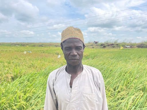A person stands in a lush green field under a partly cloudy sky, wearing a light shirt and a woven hat.