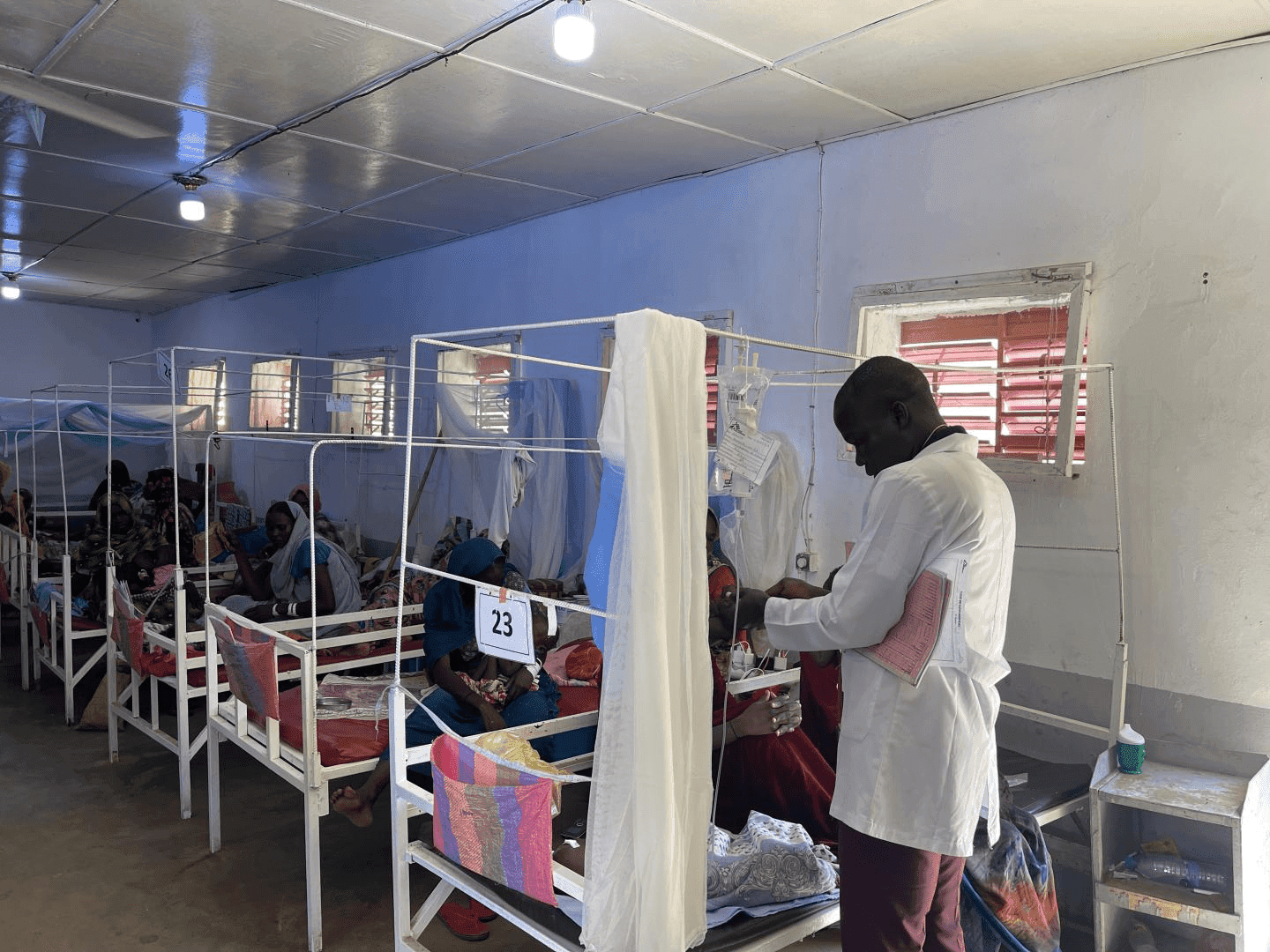 Doctor attending to a patient in a hospital ward with several beds and numbered partitions.
