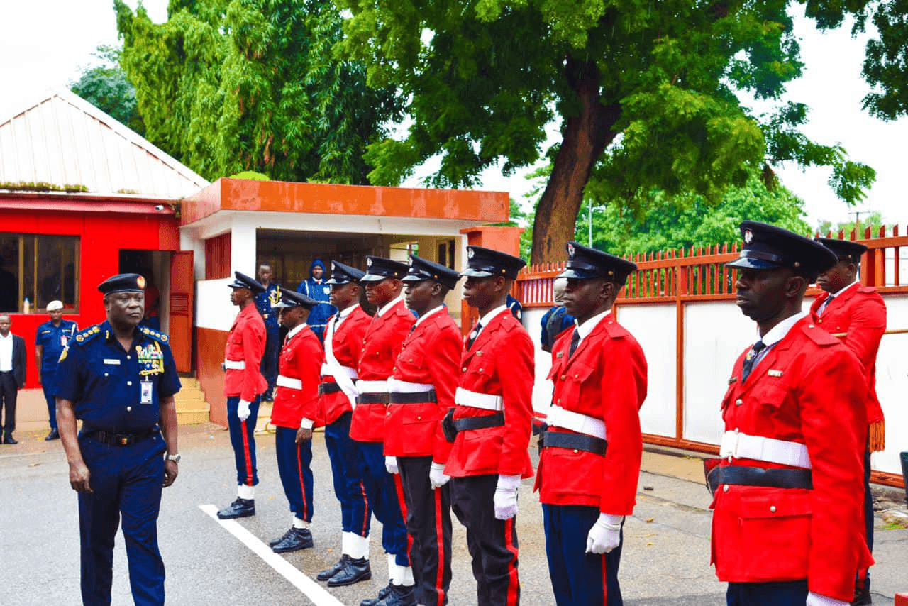 fire service in Nigeria Uniformed officers in red stand in formation, while a commanding officer in navy blue inspects them, with trees and buildings in the background.