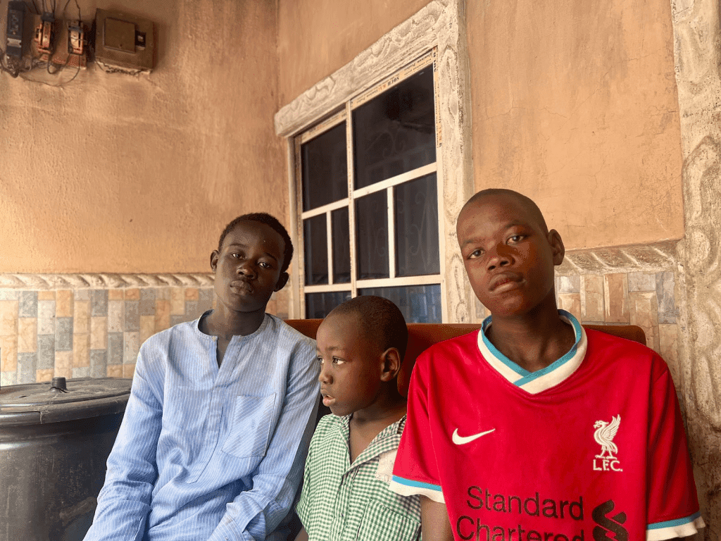 Three boys sitting indoors, one in a blue shirt, another in green, and one in a red sports jersey, looking towards the camera.