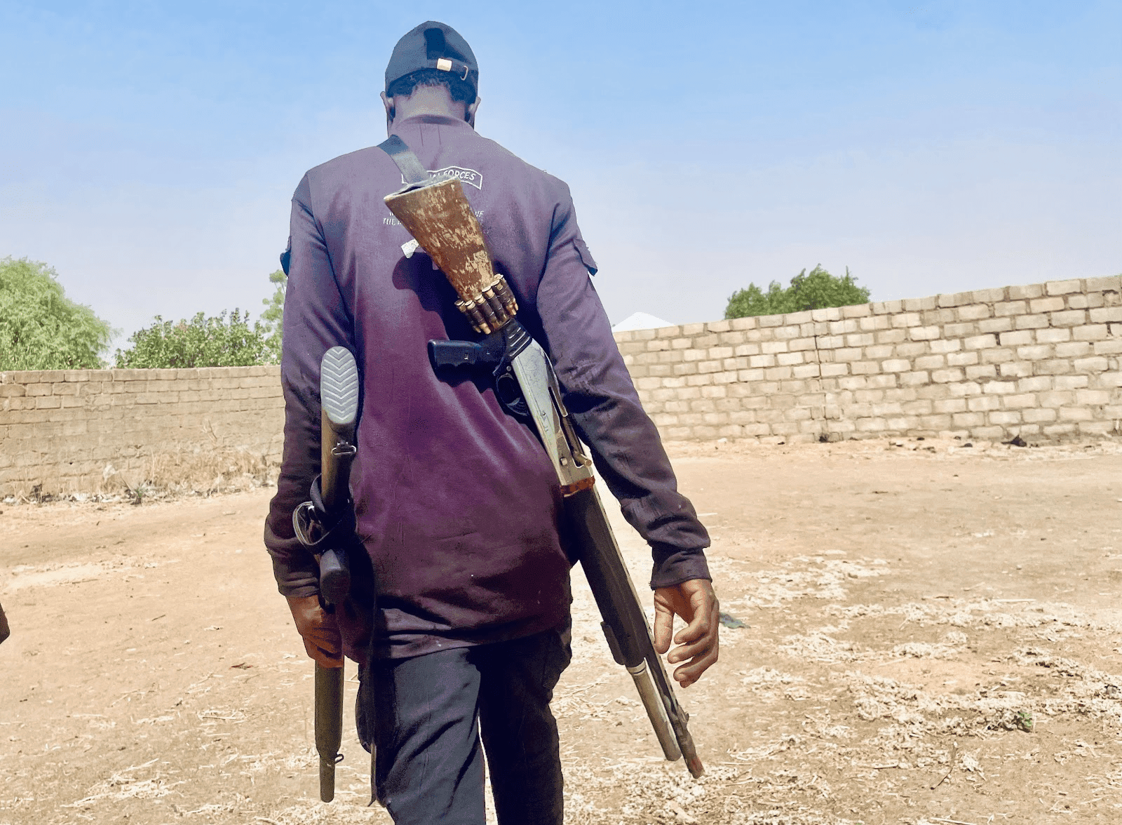 boko haram attack in lake chad Back view of a person walking outdoors with a shotgun slung over their back and holding another in hand, near a brick wall.