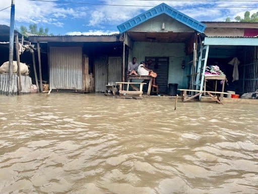 Flooded street with people sitting outside a building, surrounded by water.