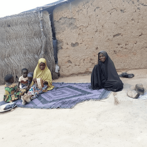 Two women and two children sit on mats outside a thatched and mud wall structure, surrounded by simple belongings.